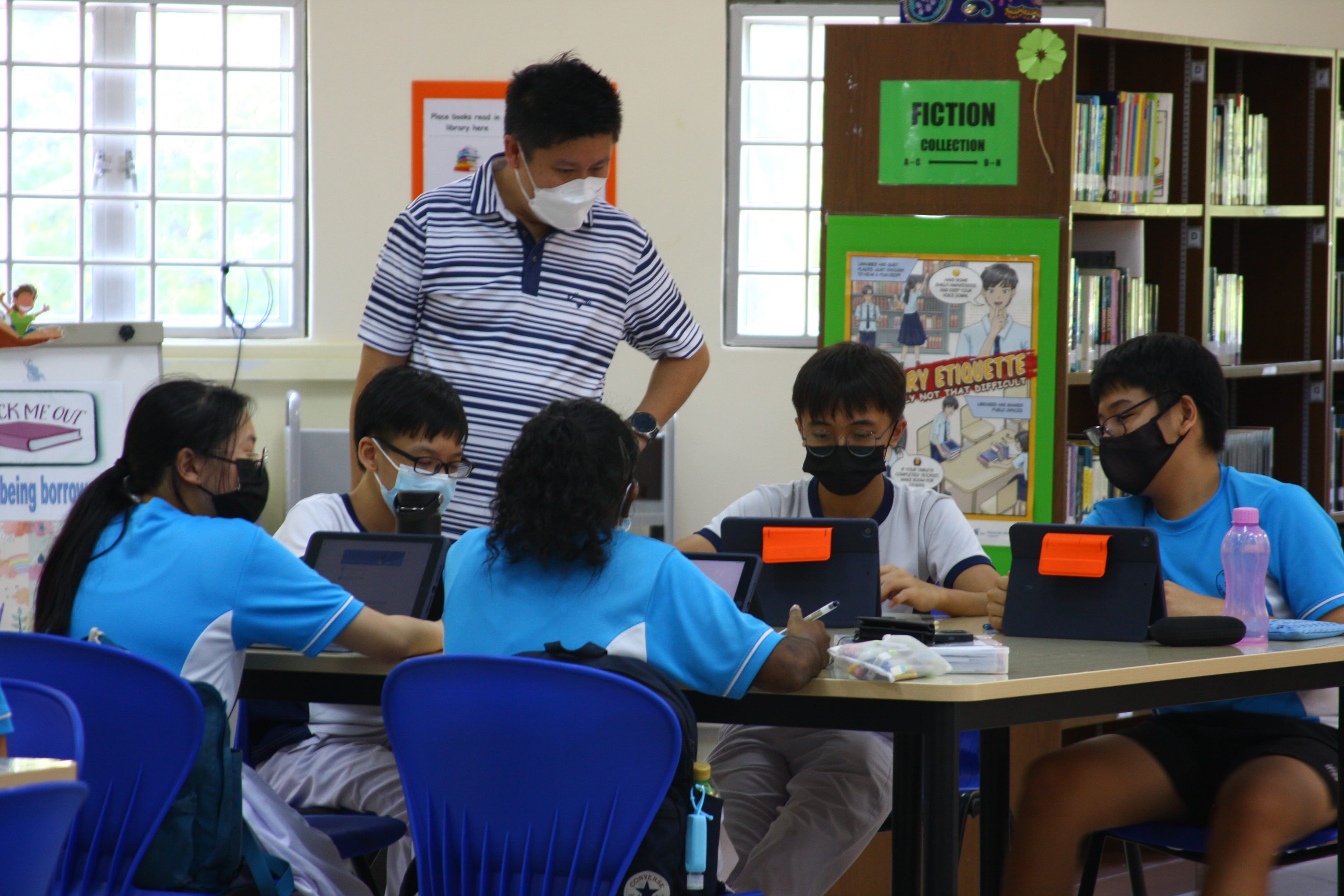 Students in blue uniforms and masks work on tablets at a library table while a masked adult supervises.